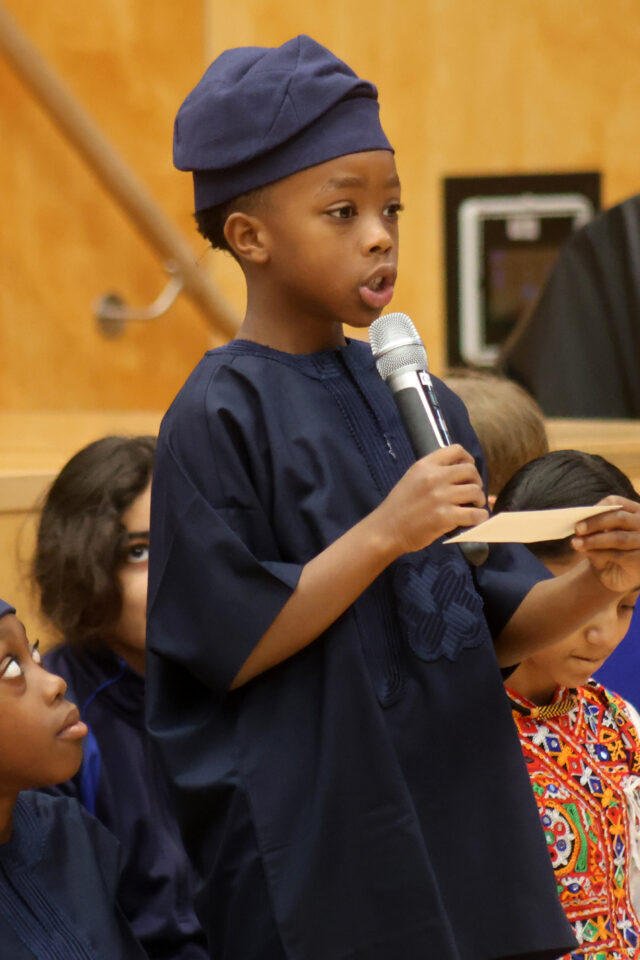 A young boy in a traditional dark blue outfit and cap speaks into a microphone whilst holding a small piece of paper.