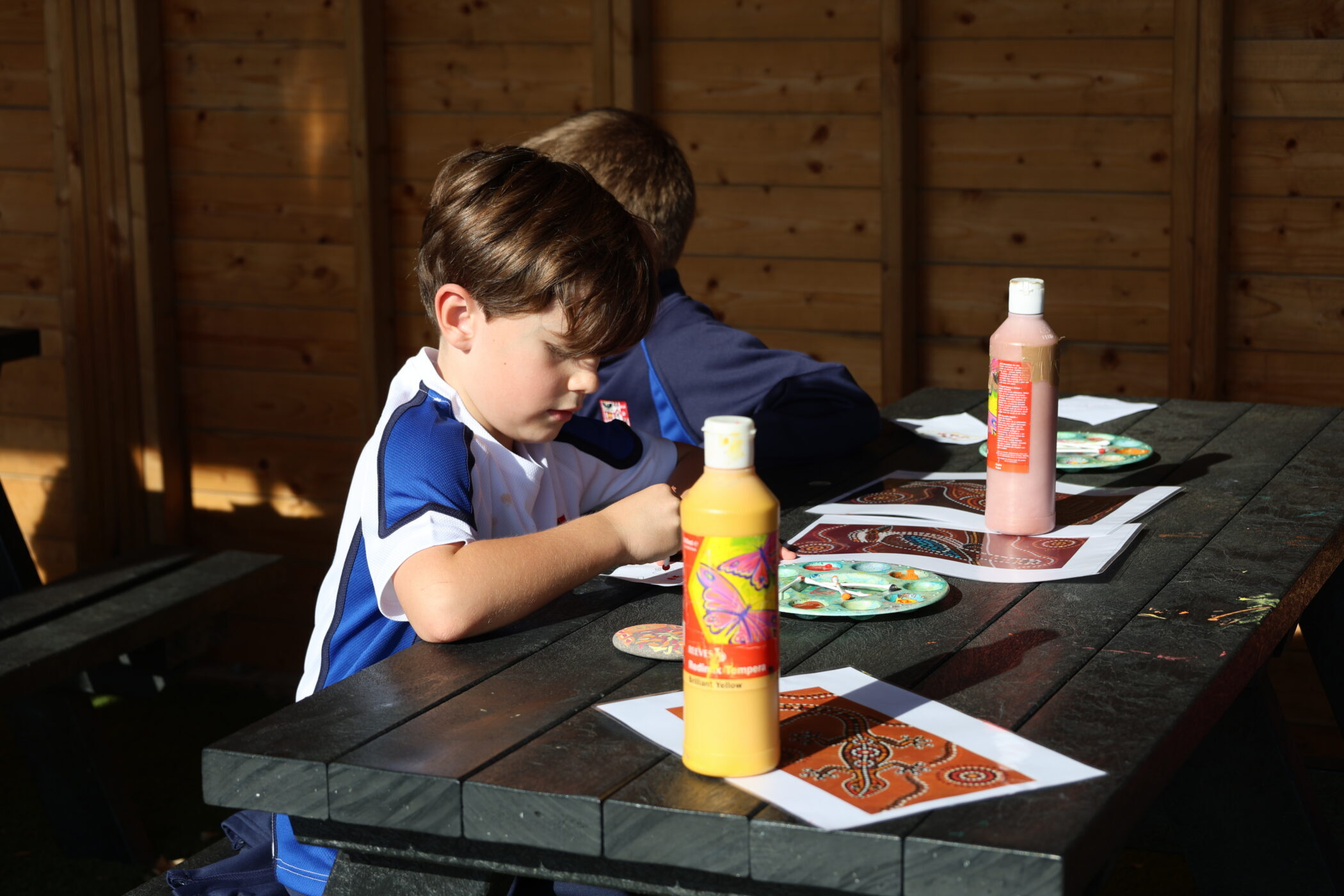 Two children sitting at an outdoor table painting artwork on paper.