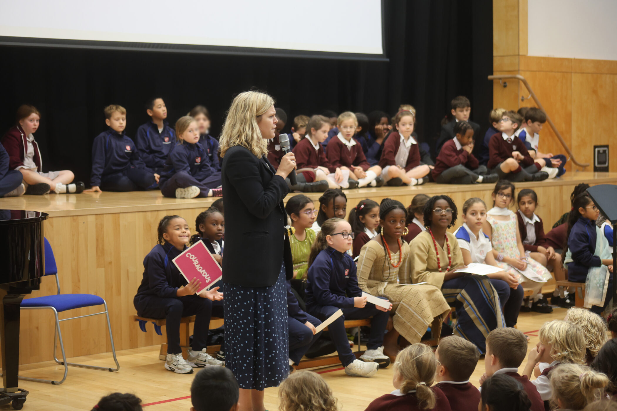 A woman in a blazer speaks into a microphone in front of an audience of students sitting on a stage and on the floor.