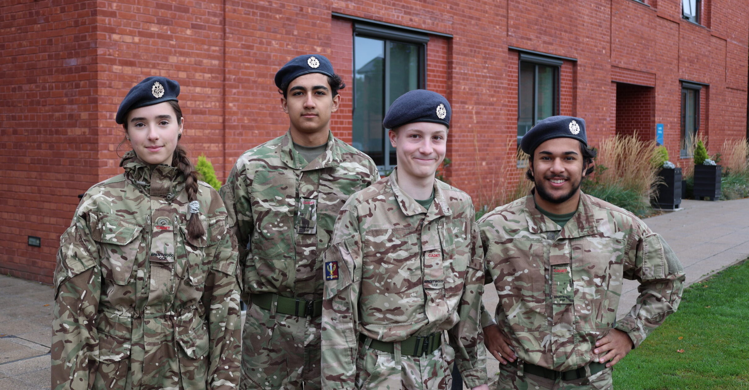 Four young co-curricular CCF cadets in camouflage uniforms and blue berets stand in front of a red brick building.