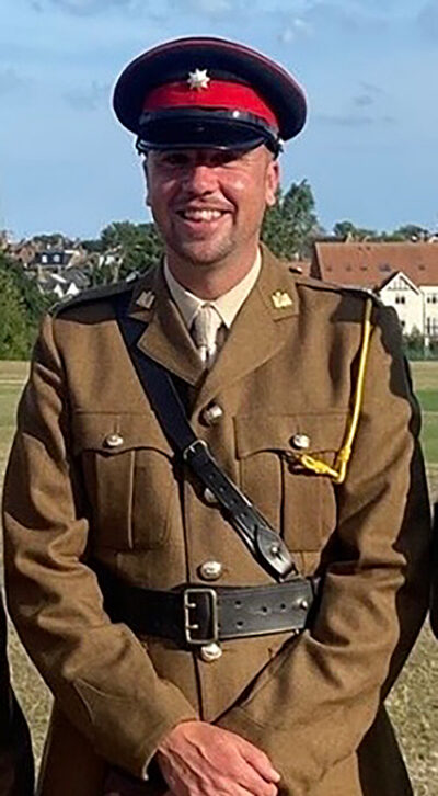 A smiling soldier in a tan dress uniform, black belt, and peaked cap with a red band.