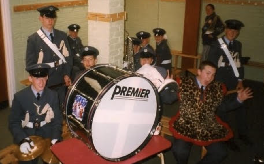 A group of band members in uniform, including one person in a leopard-print tunic, pose with a large Premier bass drum.