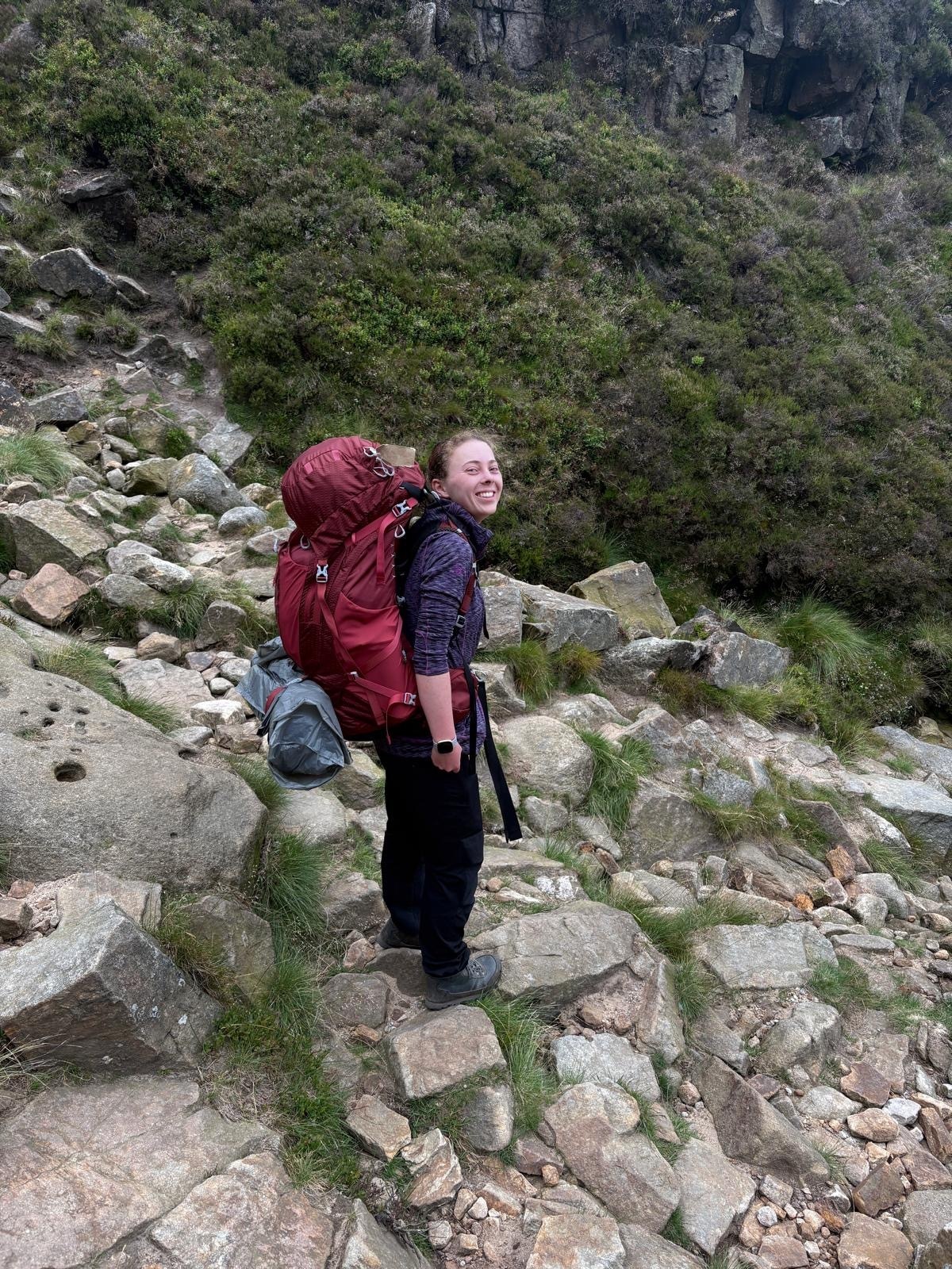 A smiling woman with a large red rucksack stands on a rocky co-curricular footpath.