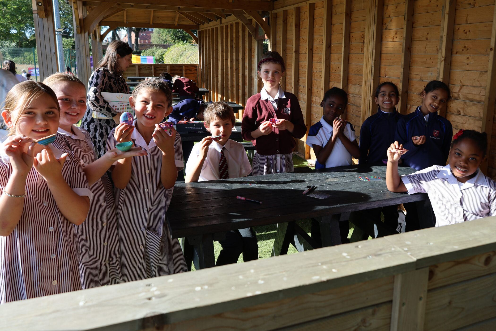 A group of schoolchildren in uniforms sitting at picnic tables outdoors, some holding small painted items.
