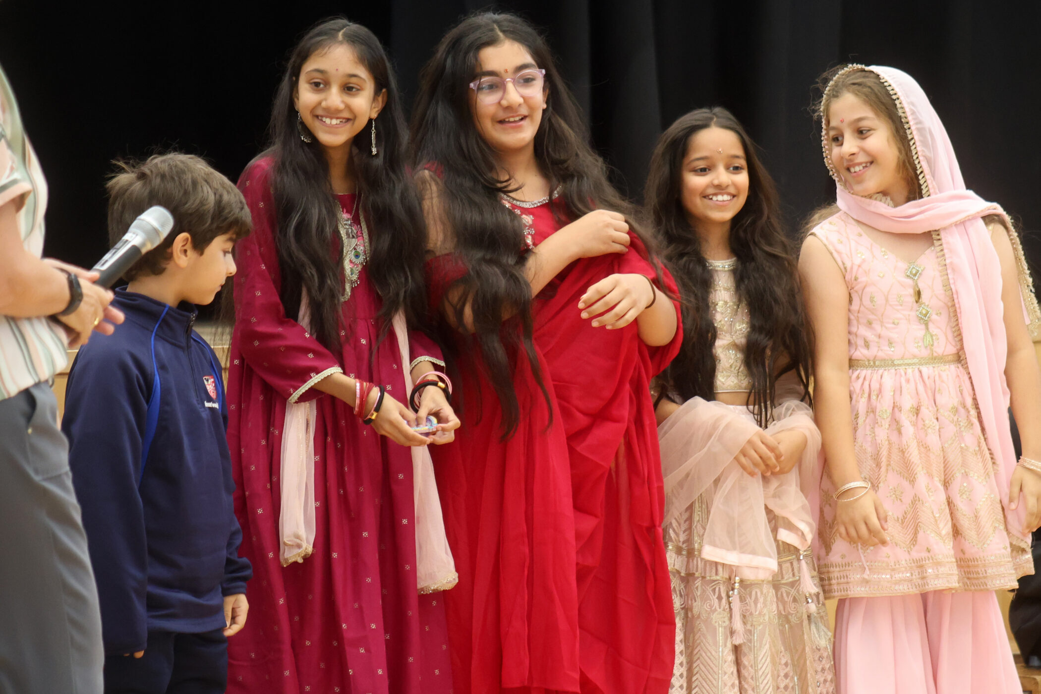 Five children in traditional South Asian attire stand together on stage, smiling and laughing. One adult is partially visible holding a microphone on the left side. The background is dark and out of focus.