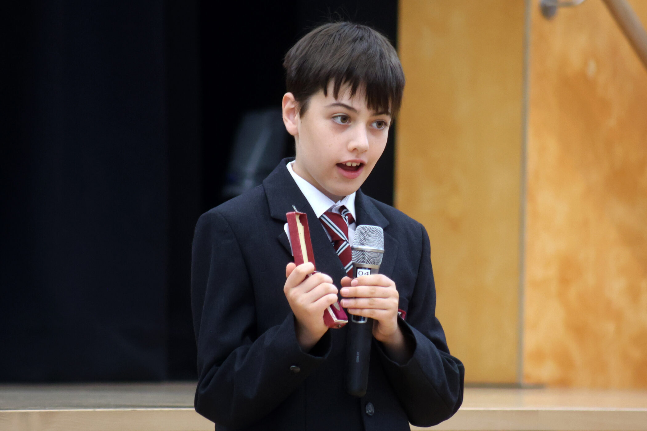A young boy in a suit and striped tie holds a microphone in one hand and a small rectangular object in the other whilst speaking indoors.