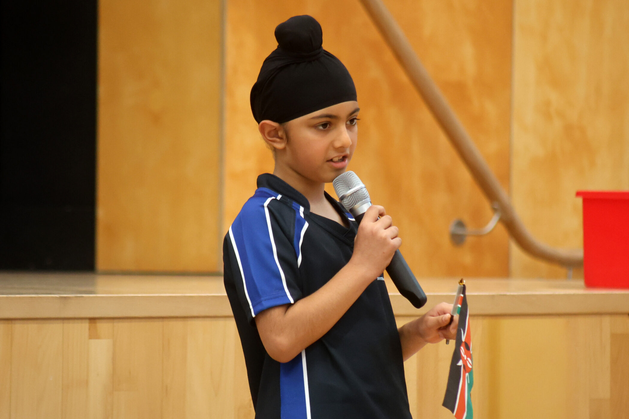 A young boy in a black turban and sports shirt speaks into a microphone while holding a small flag, standing indoors near a stage with a wooden background.