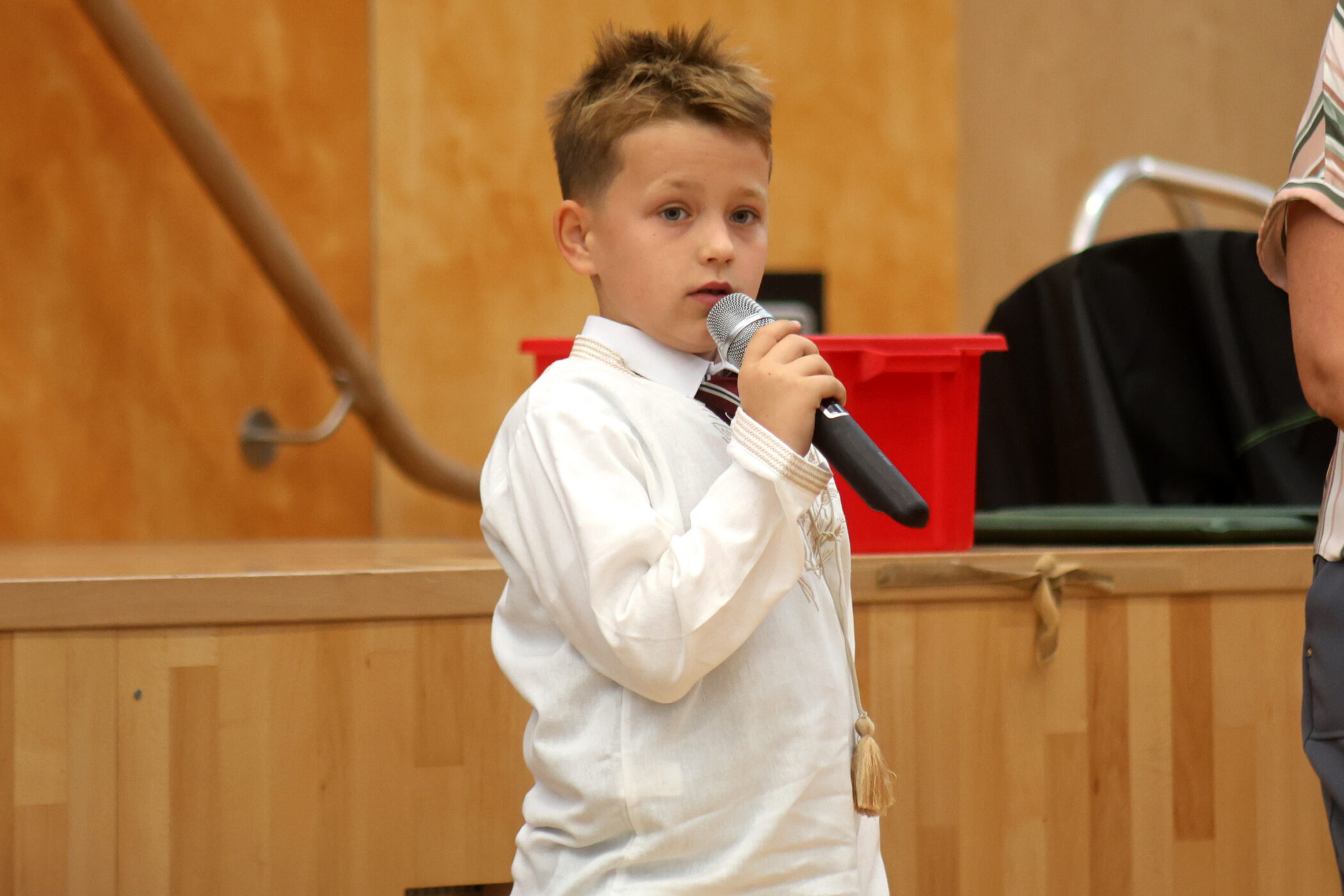 A young boy in a white shirt holds a microphone whilst speaking or singing indoors. A red bin and a wooden stage are visible in the background.