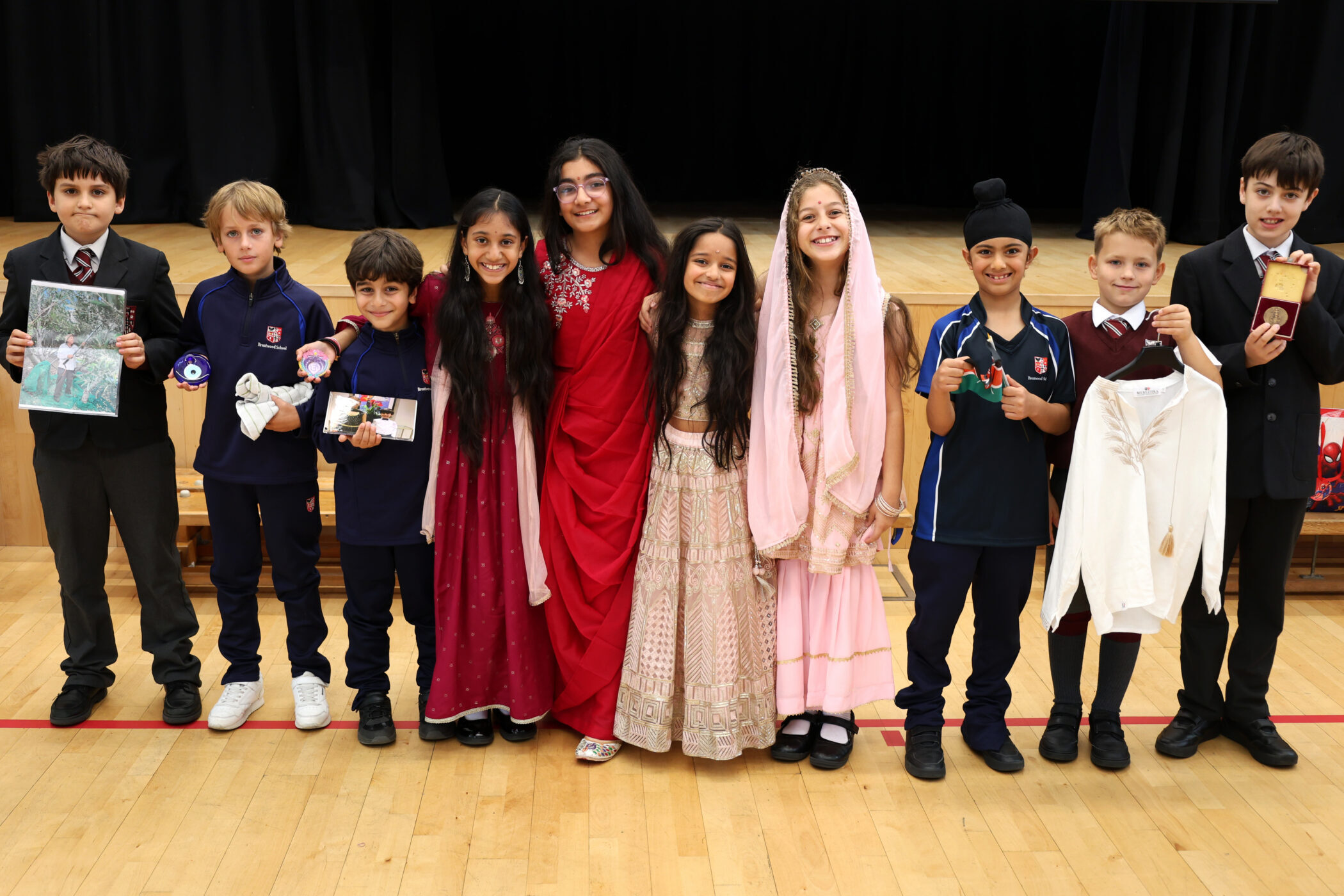 Ten children stand in a row on a gym floor, some dressed in cultural attire and holding objects, smiling at the camera with a black curtain in the background.