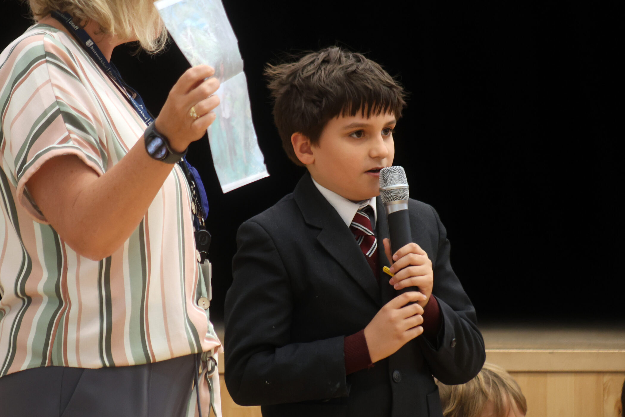 A young boy in a suit speaks into a microphone while a woman beside him holds up a clear plastic sheet. They appear to be on a stage or at an event.