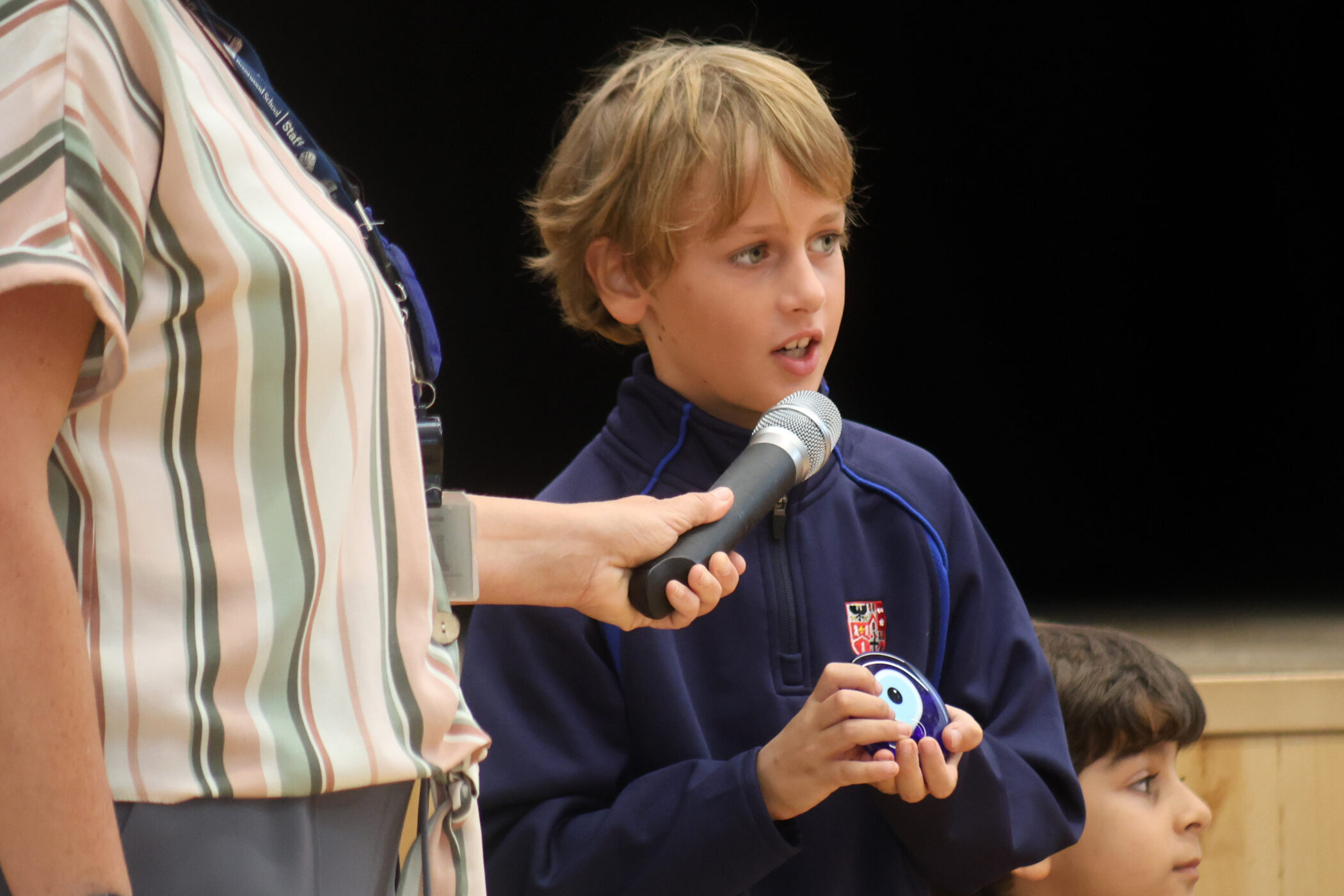 A young boy speaks into a microphone held by an adult whilst holding a small round object, with another child sitting nearby.