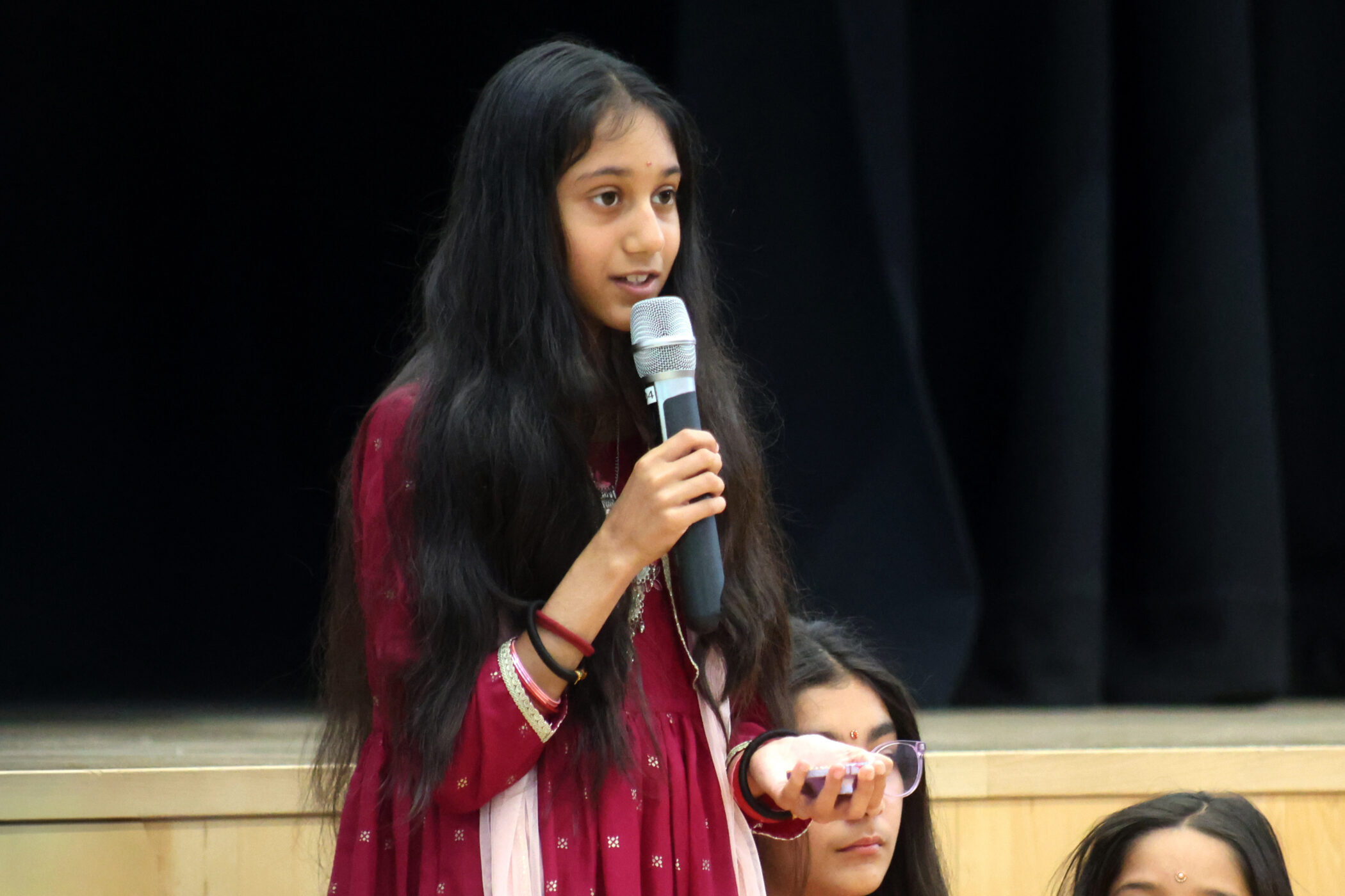 A young girl with long dark hair, wearing a red traditional outfit, speaks into a microphone whilst standing on a stage with a dark curtain in the background.