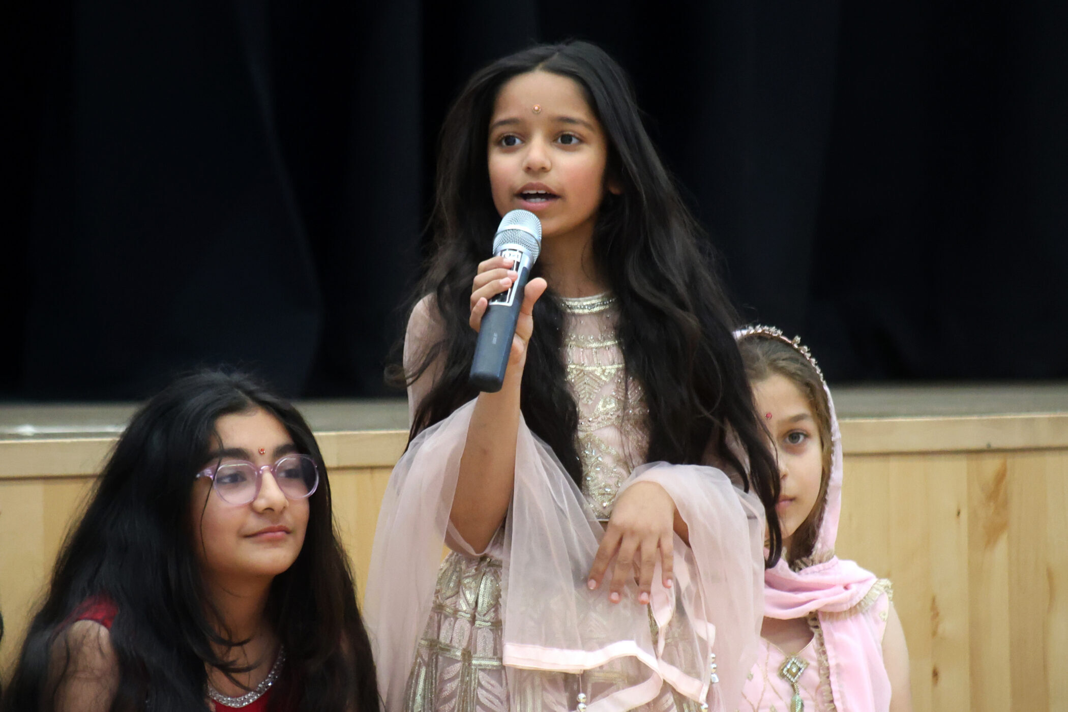 A girl in traditional attire holds a microphone and speaks, whilst two other girls sit beside her, also dressed in traditional clothes, against a stage backdrop.