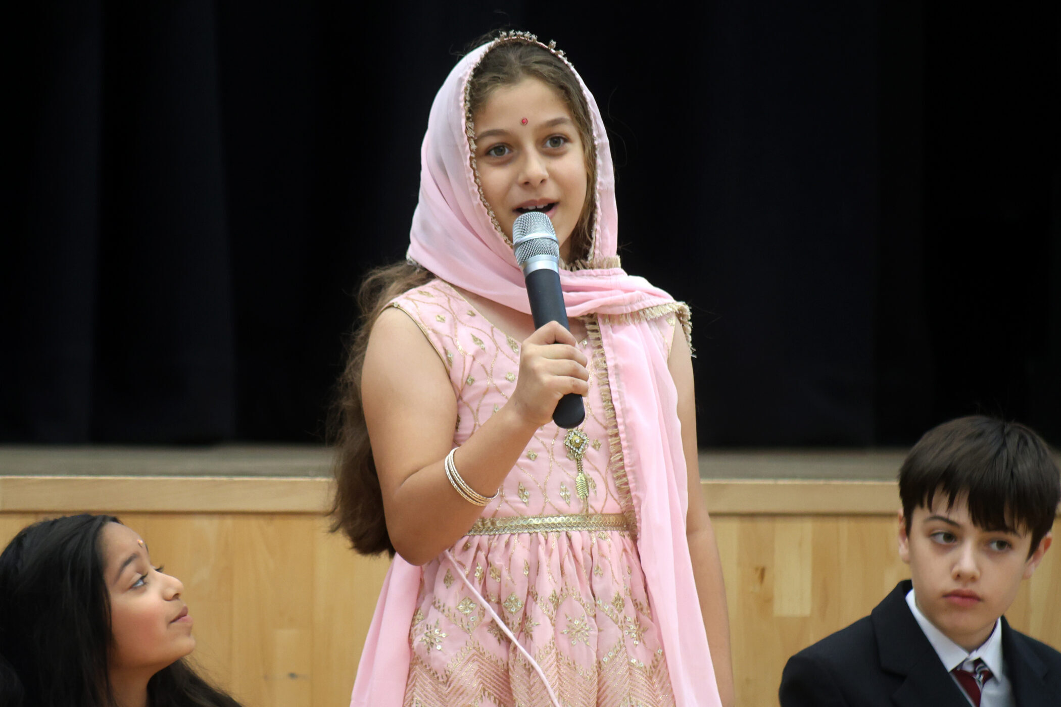 A girl in a pink traditional outfit and headscarf speaks into a microphone on stage, whilst two other children sit nearby and listen attentively.