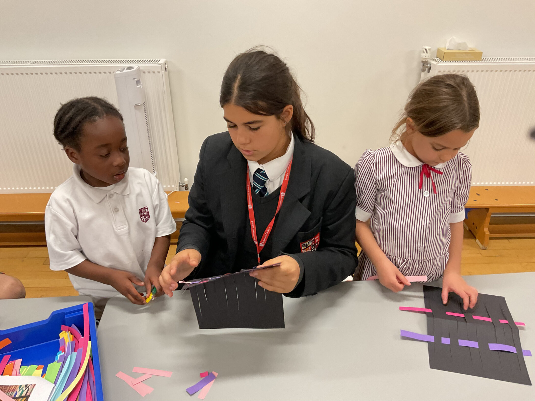 Three students in school uniforms working on a paper weaving craft project at a table.