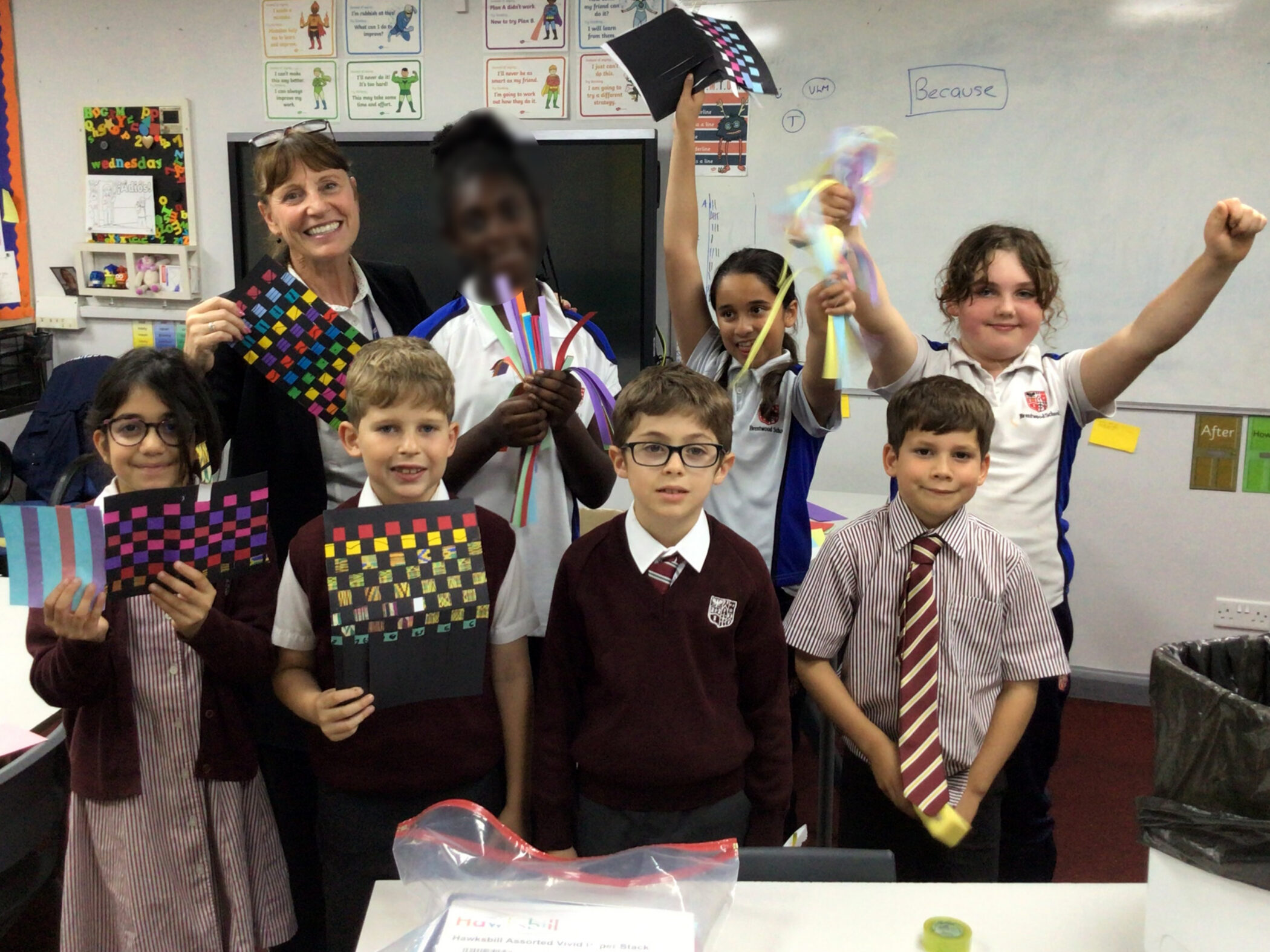 A teacher and seven primary school pupils in uniform hold up their colourful paper weaving projects in a classroom.