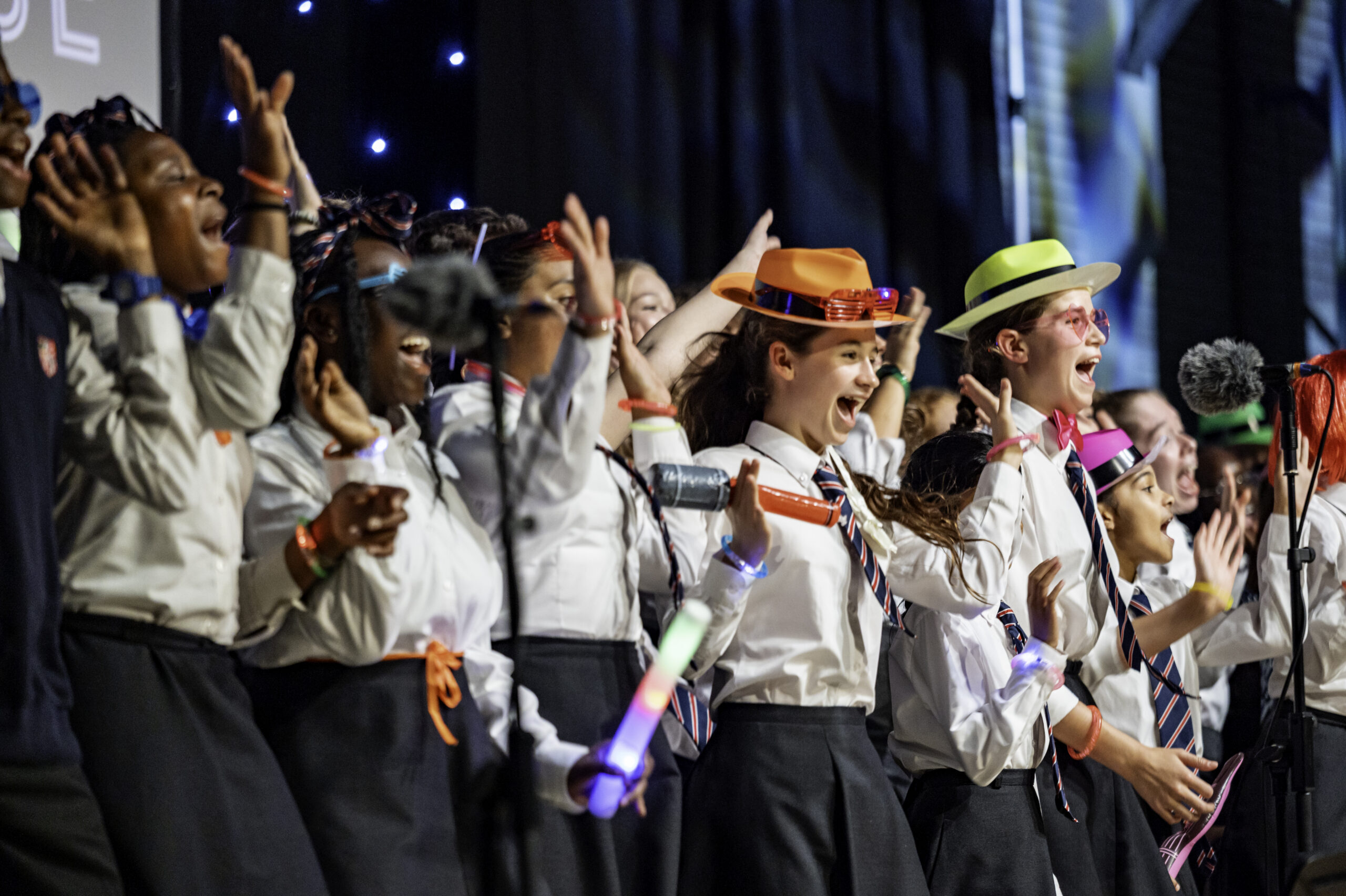 A group of students in uniforms enthusiastically sing on stage, some wearing colourful hats and holding glow sticks, showcasing how music education builds confidence and communication skills.
