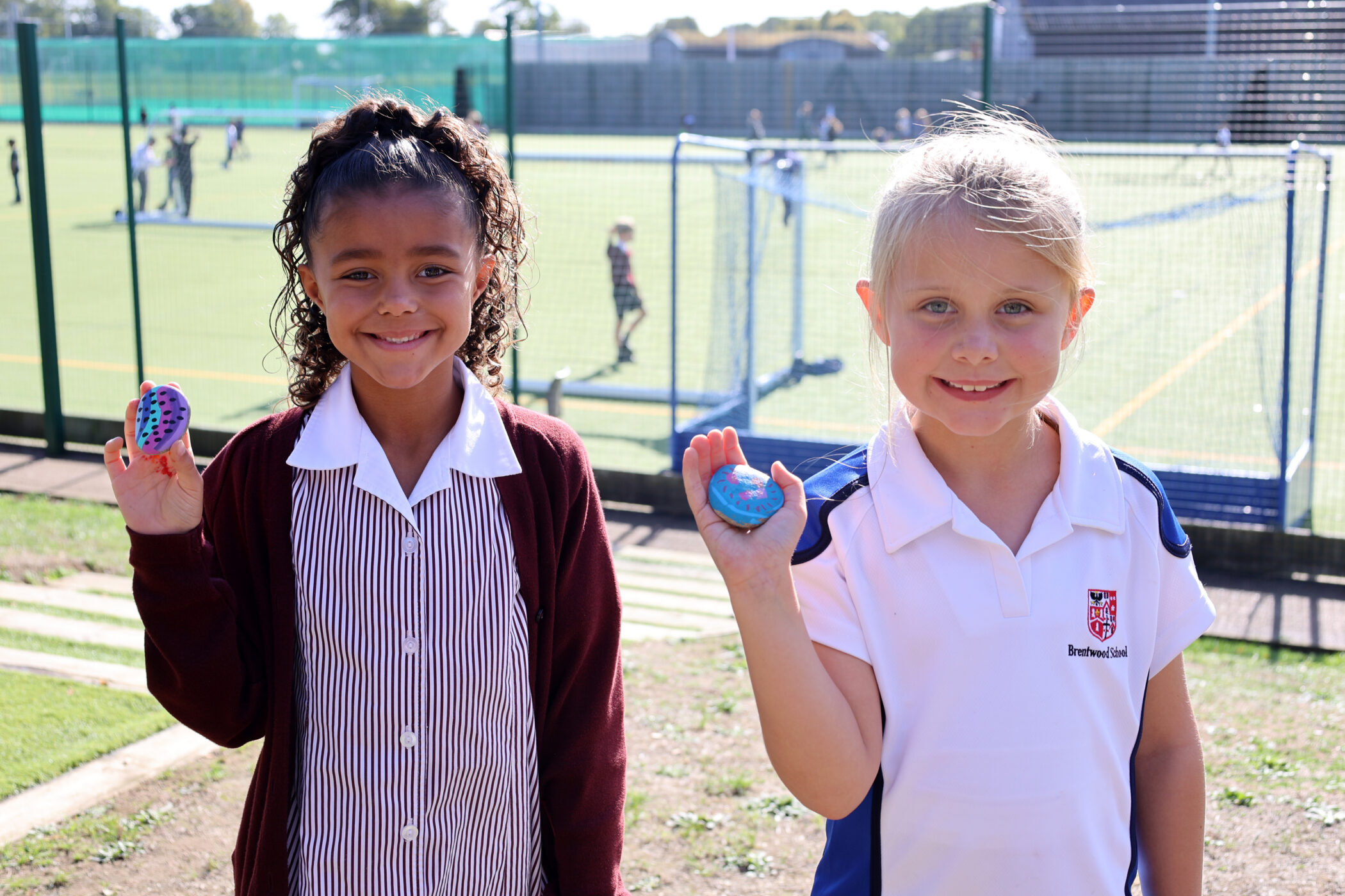 Two schoolgirls smiling whilst holding up painted stones on a sports field.