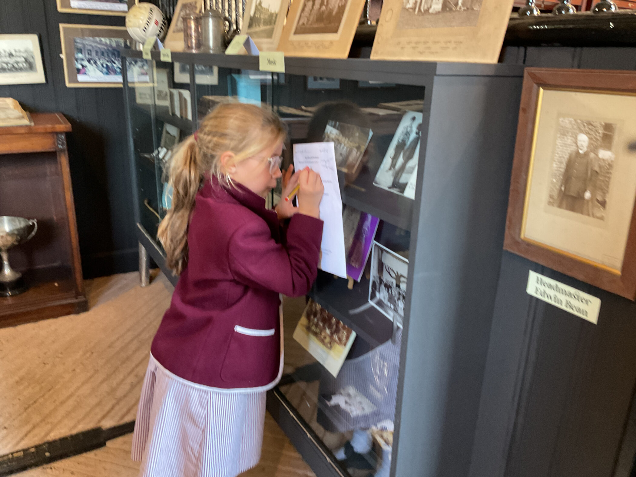 A young girl in a school uniform writes on a piece of paper held against the glass of a museum display case filled with historical photographs and artefacts.