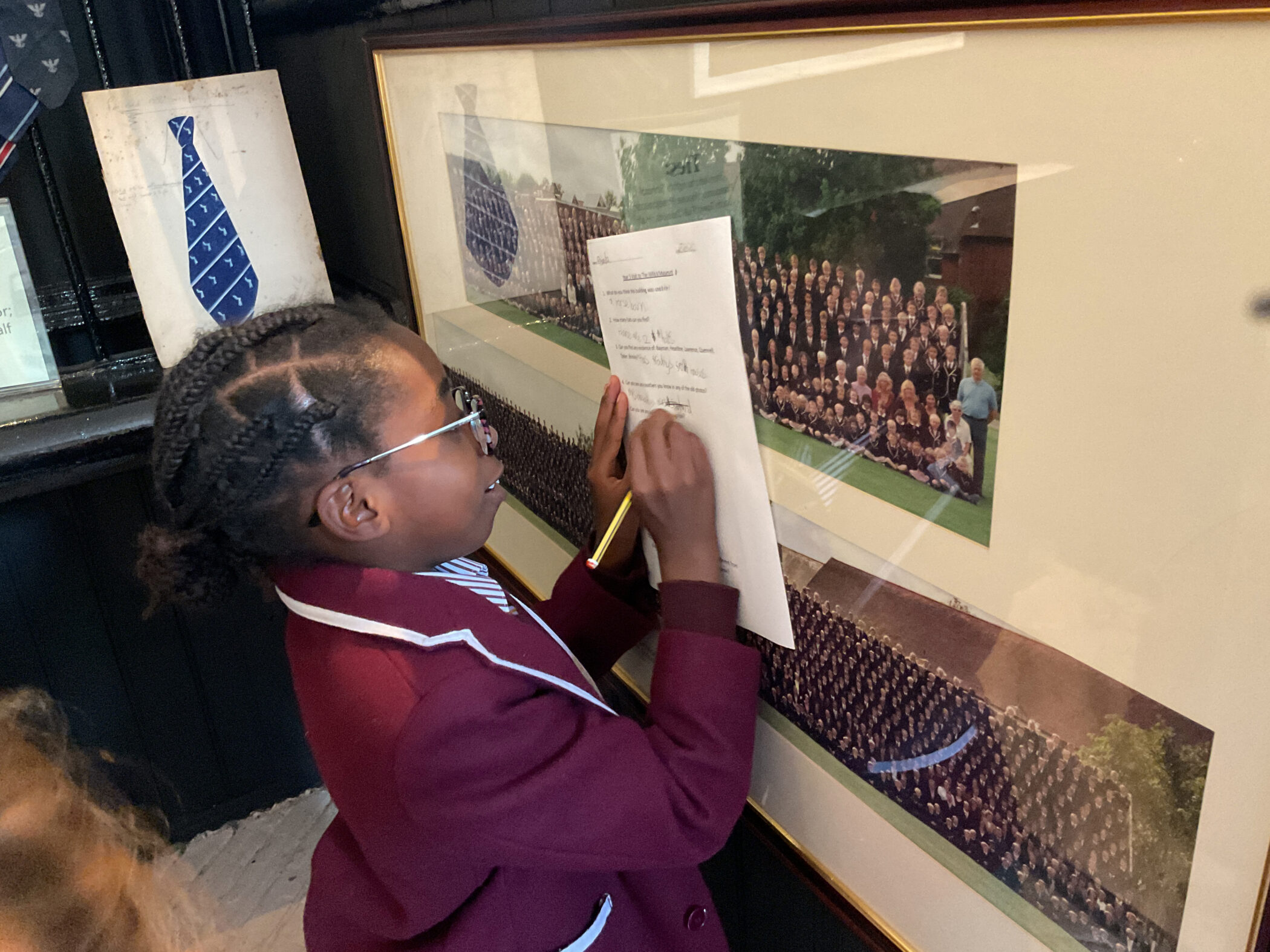 A student in a maroon school blazer writes on a piece of paper held against a large framed photograph of a group of people.