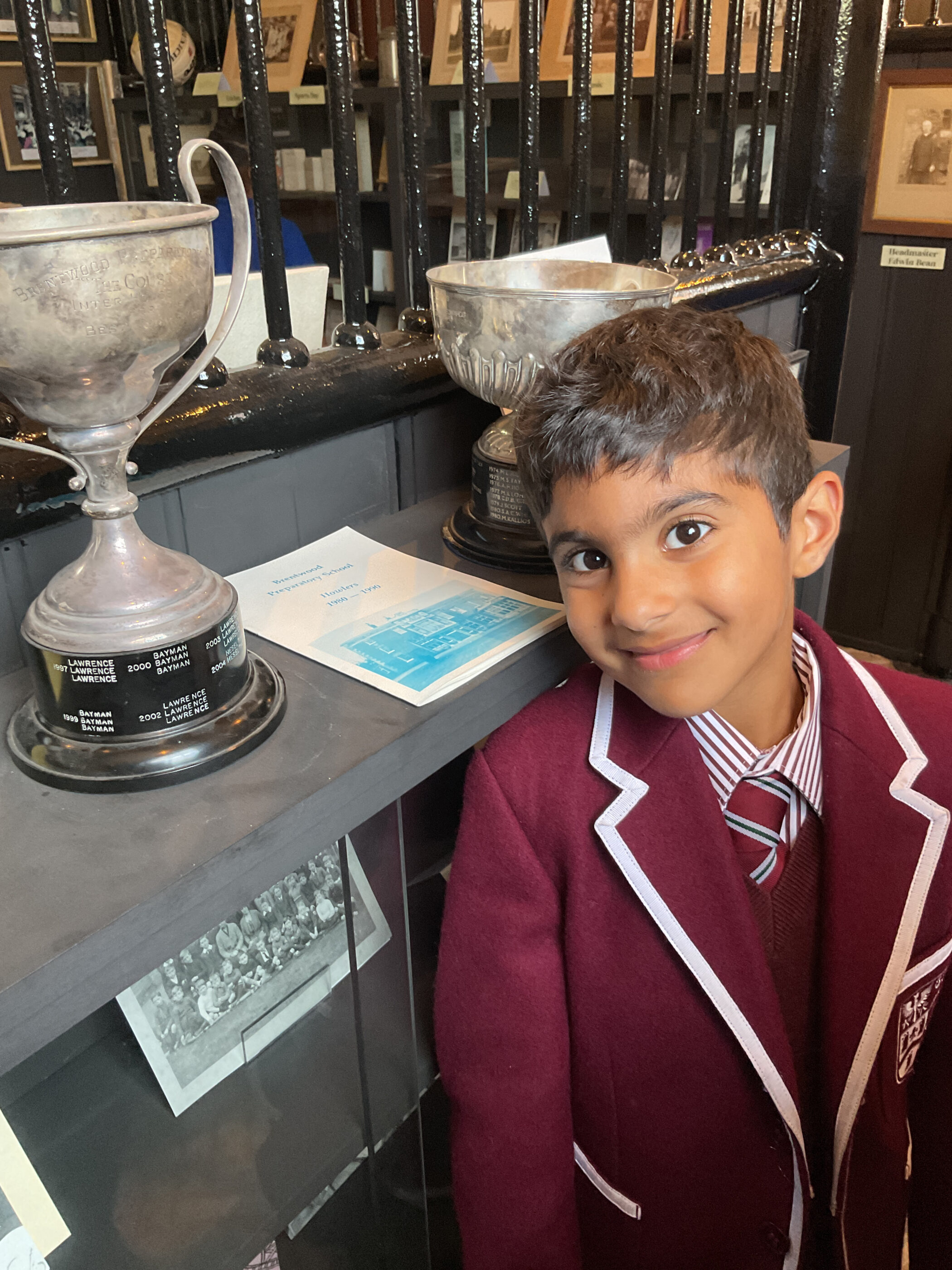 A schoolboy in a maroon blazer and striped tie smiles next to a trophy on a display shelf.