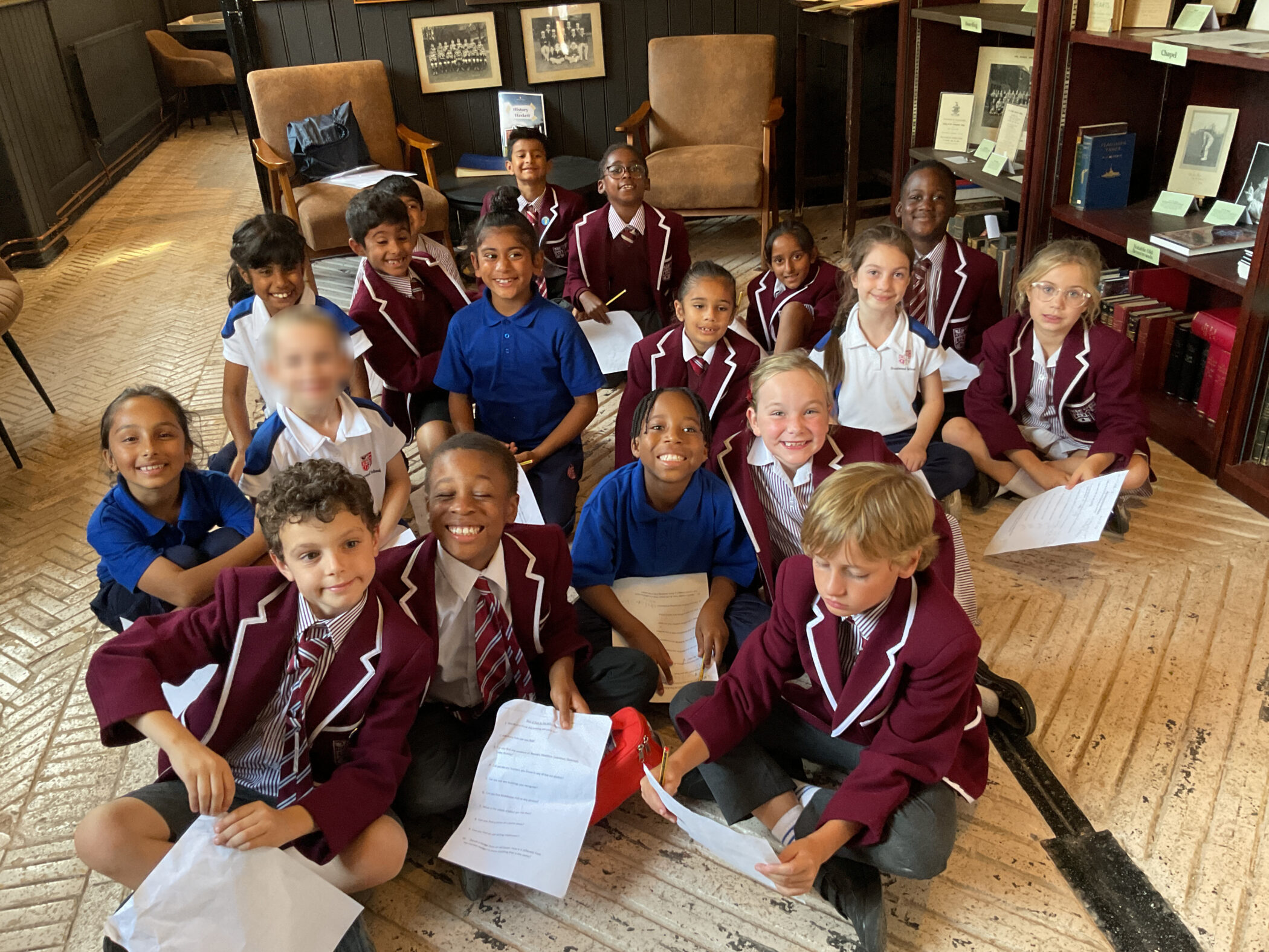 A group of students in school uniforms sitting on the floor in a library, holding sheets of paper.
