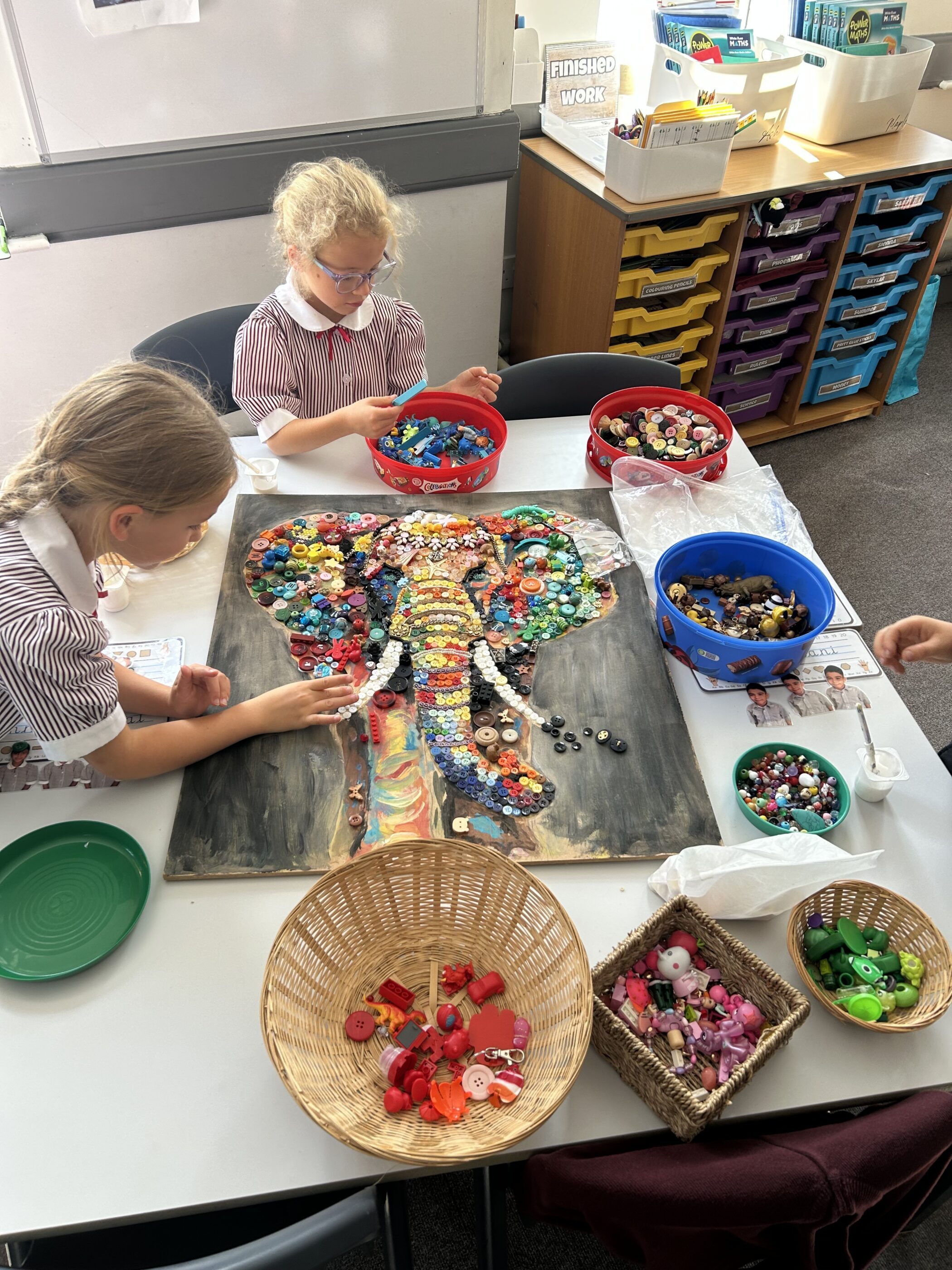 Two girls in school uniforms create an elephant mosaic using colourful buttons on a classroom table surrounded by containers of supplies.