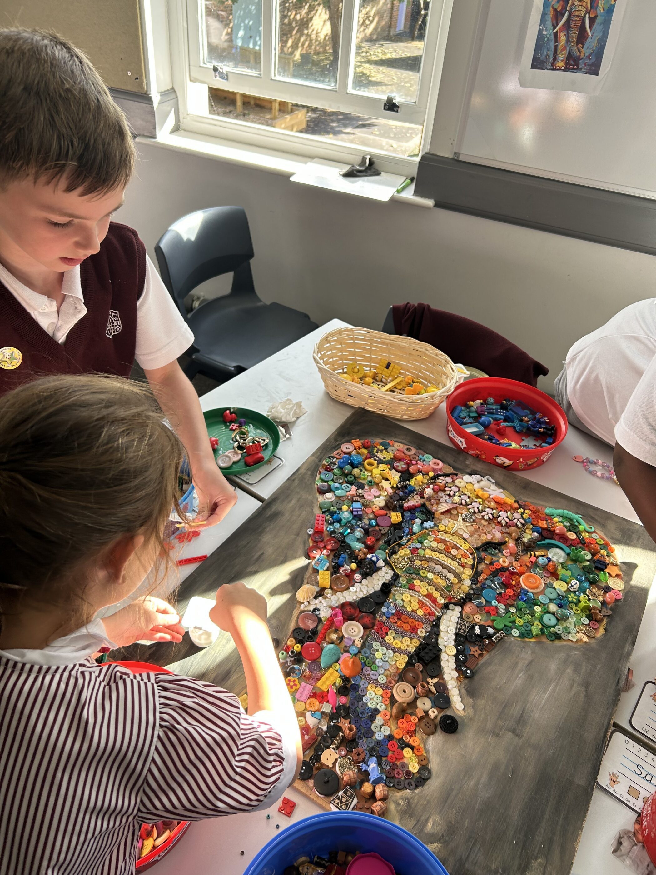 Two students work together to create a mosaic of an elephant made from colourful buttons and beads on a wooden board.
