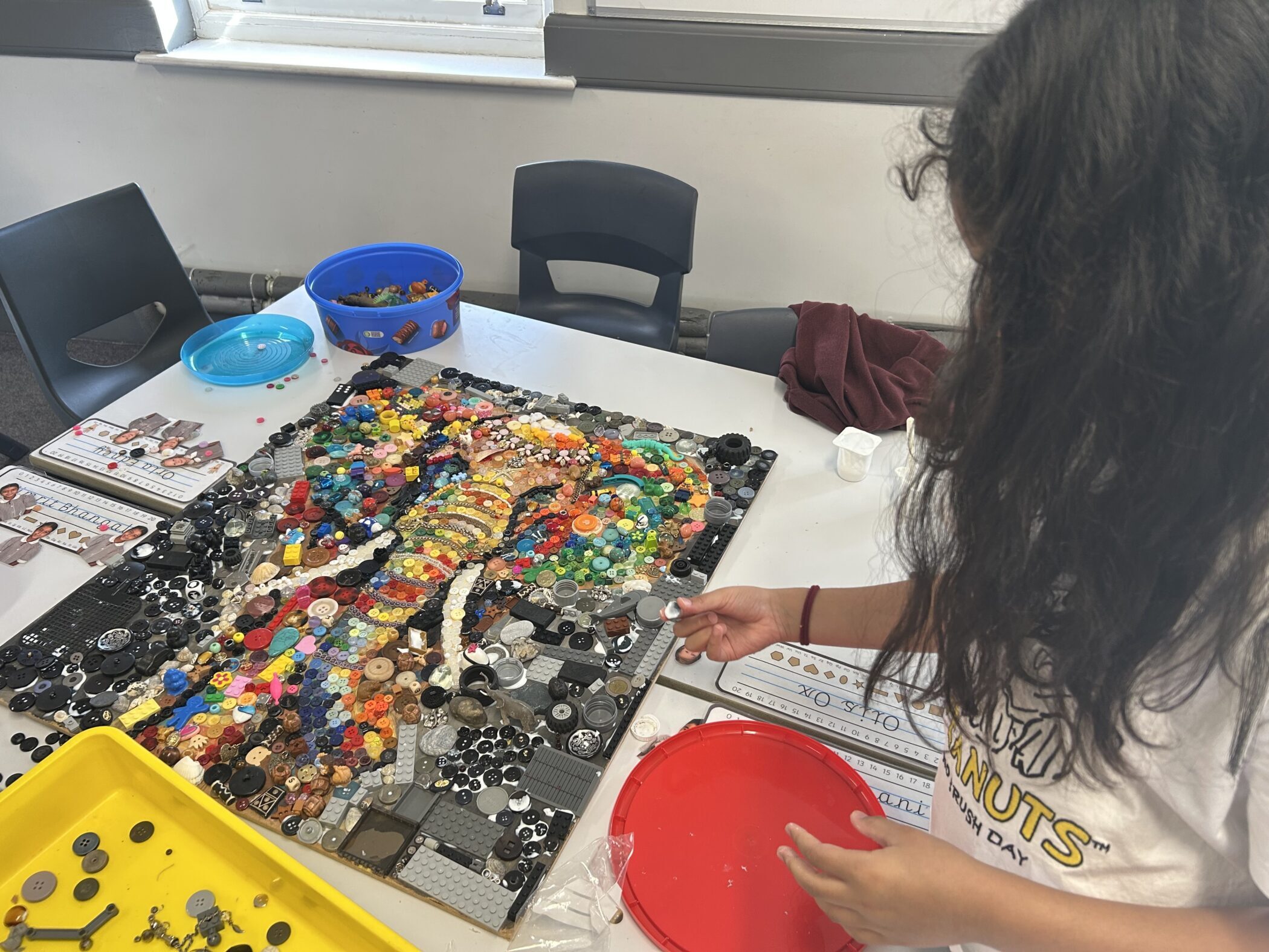 A person assembling a large mosaic of a colourful elephant made from buttons, beads, and toy parts on a table.