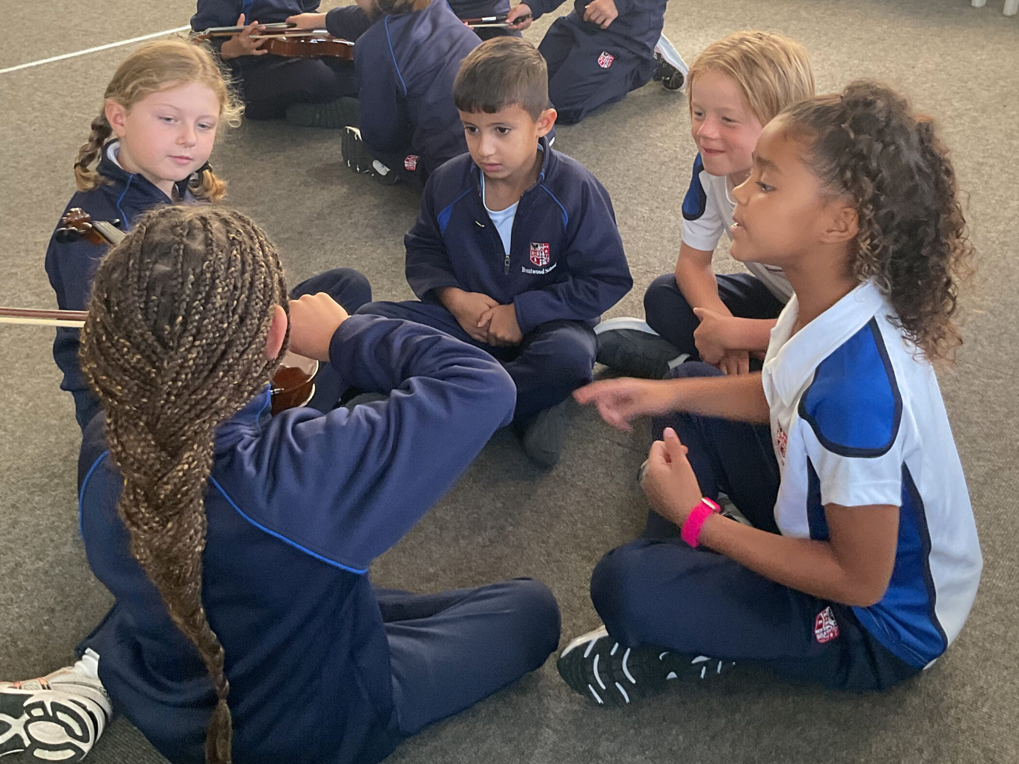 Five children in school uniforms sit in a circle on the floor, one holding a violin whilst another gestures during a lesson.