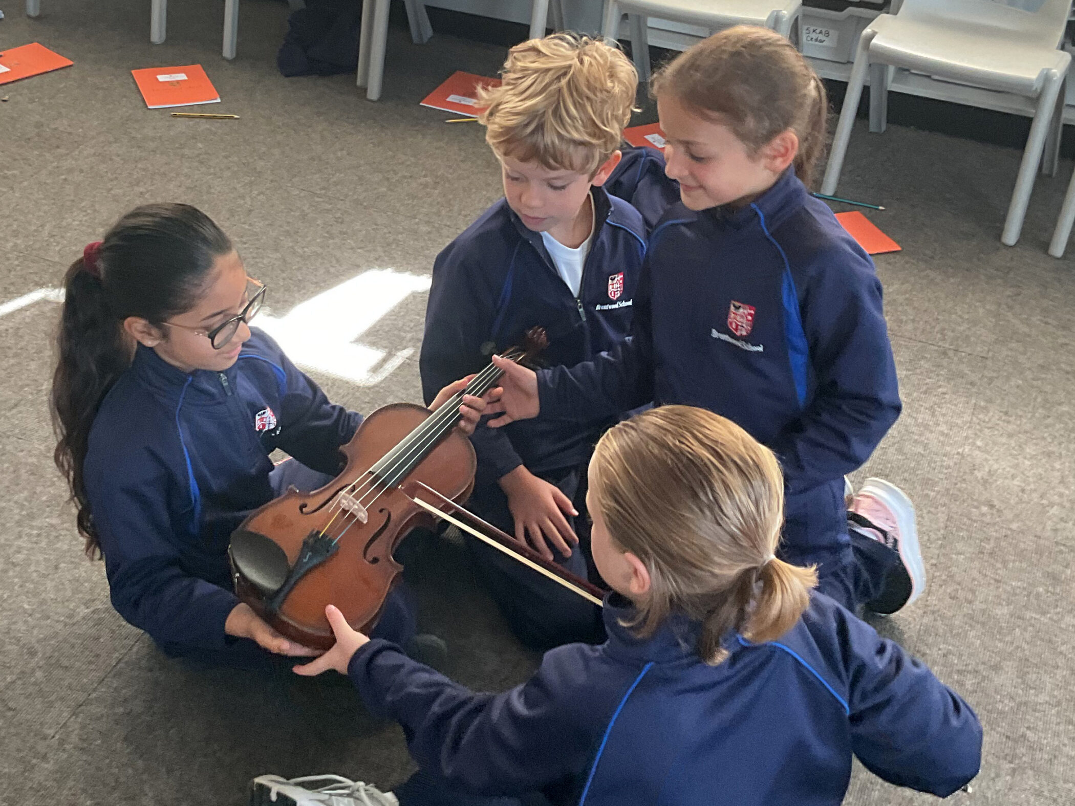 Four students in matching school uniforms sit on a carpet, looking at a violin one pupil is holding.