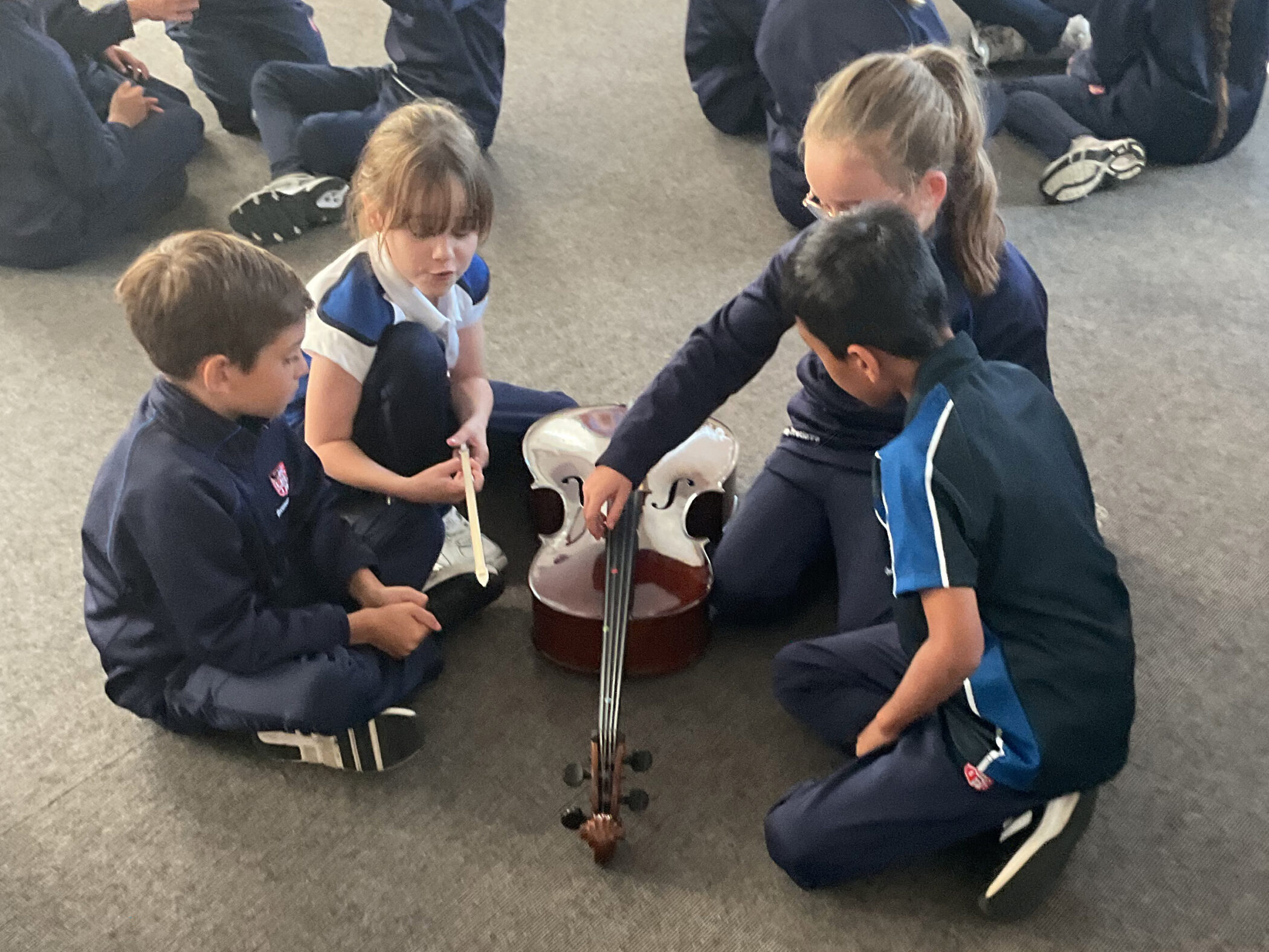 Four students in school uniforms sitting on the floor in a circle around a violoncello.