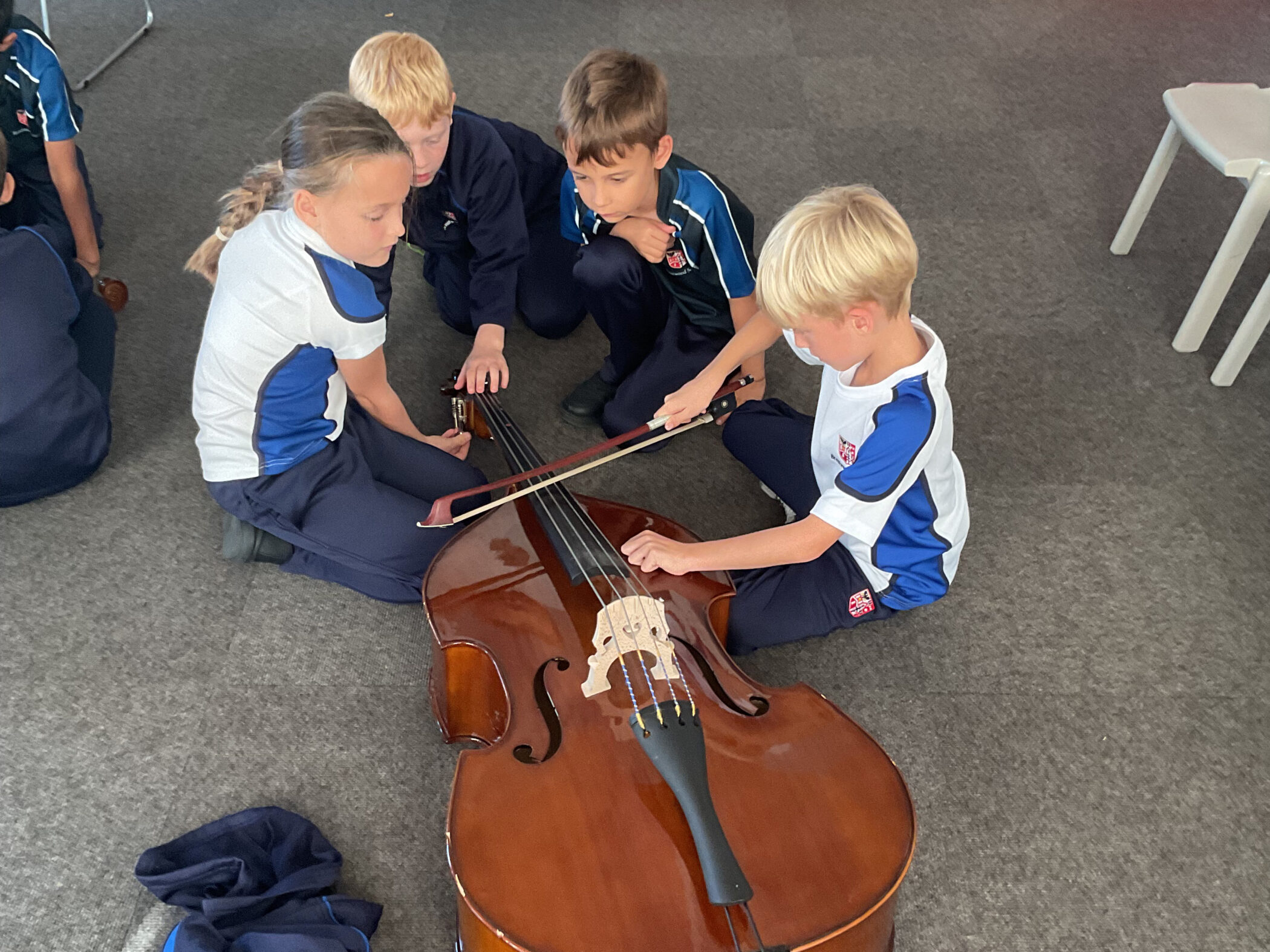 Four children in school uniforms sit on the floor, gathering round and playing a double bass.