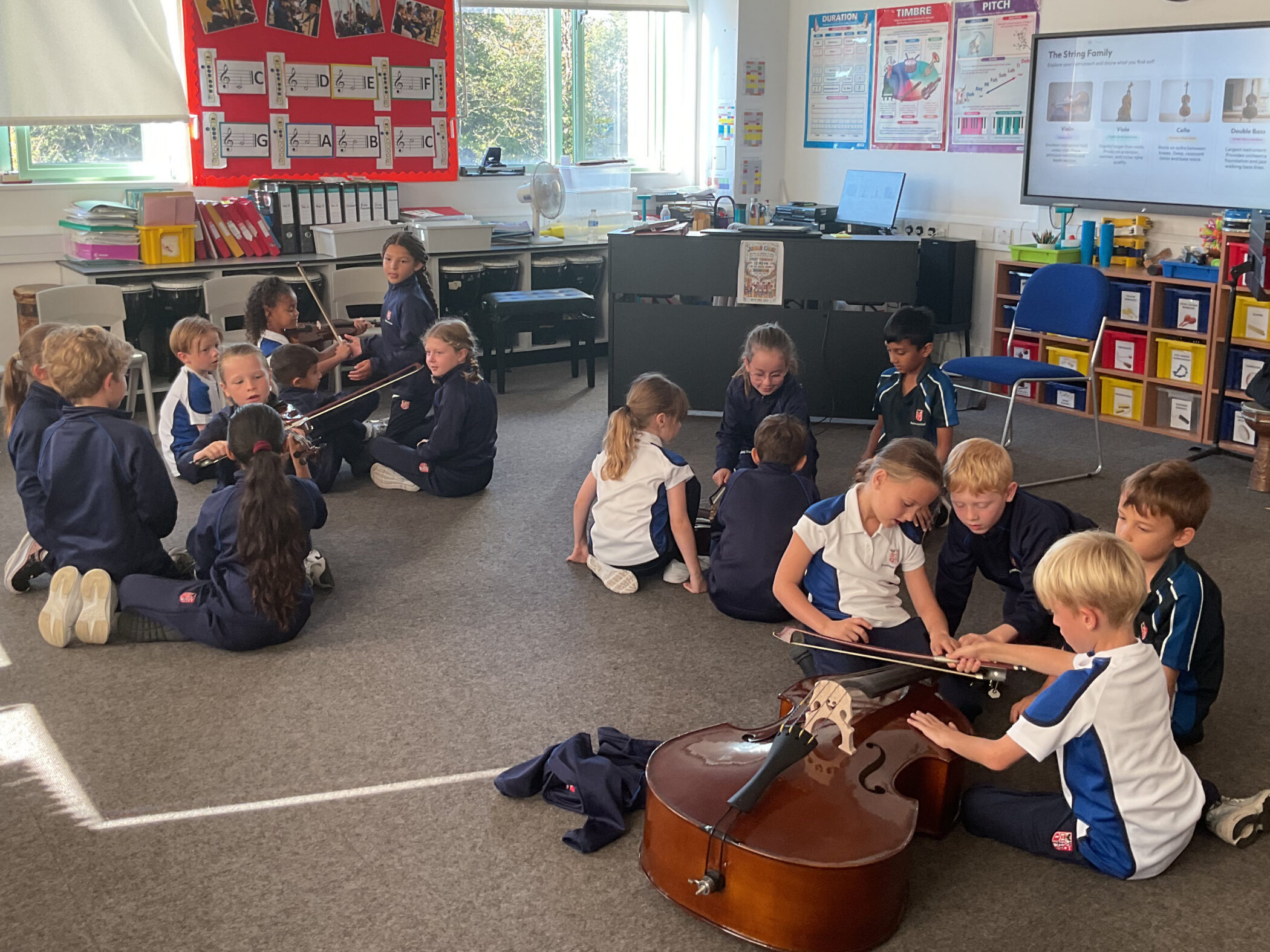 A classroom of children in school uniforms practise playing violins and a double bass on the floor.