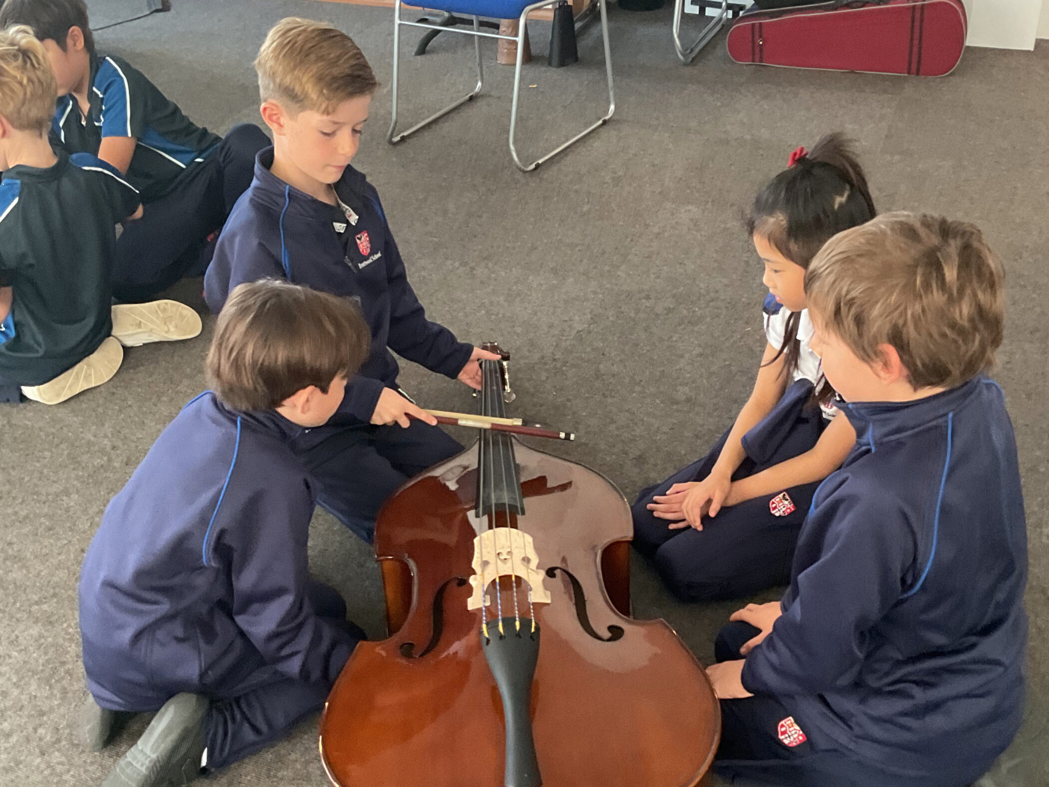 Four students in matching school uniforms sit on the floor around a double bass, watching as one boy demonstrates how to hold the bow.