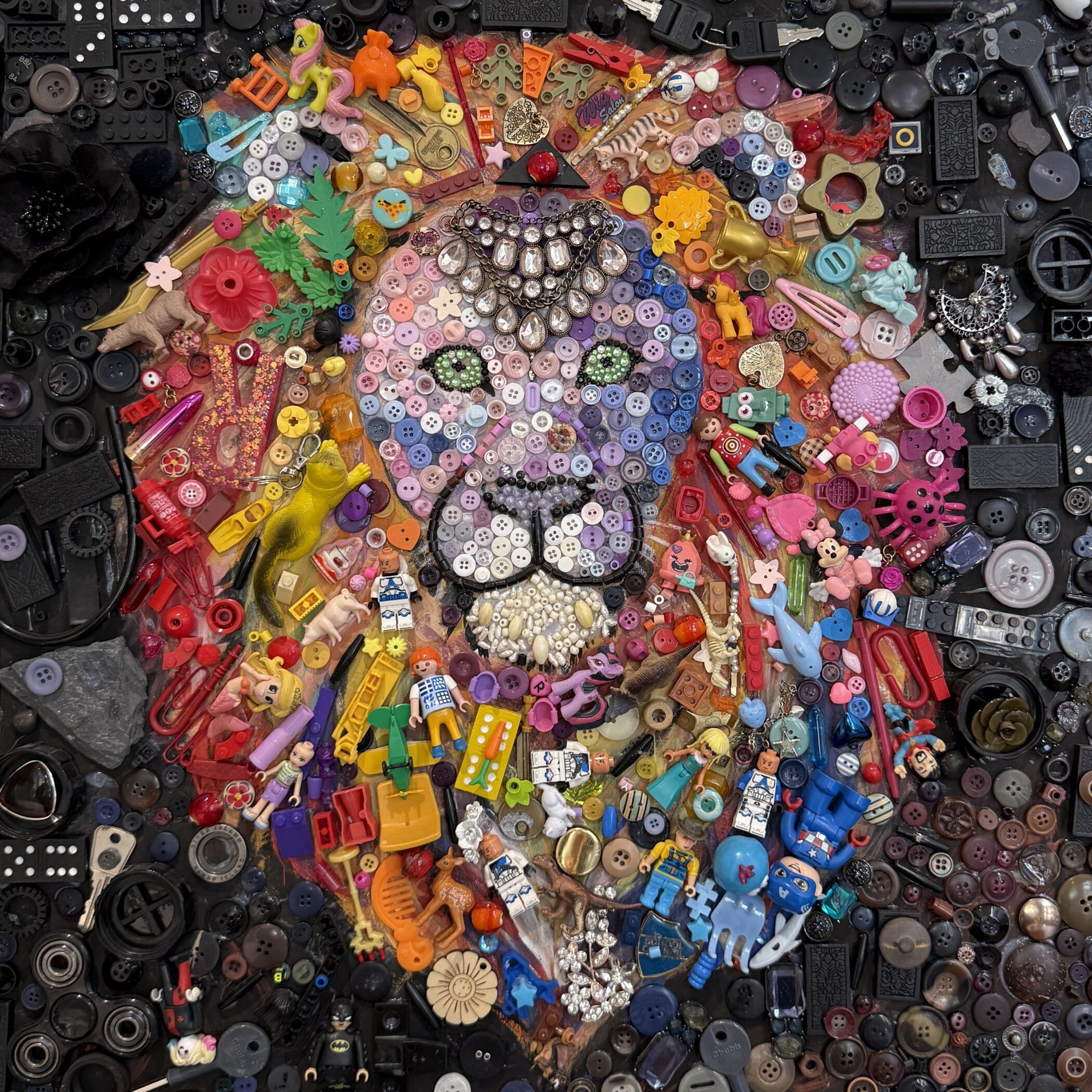 A mosaic of a lion's face created from assorted colourful buttons, plastic toys, jewellery, and small trinkets, set against a dark background of buttons and hardware.
