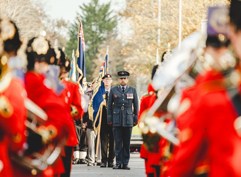 A military officer in uniform standing in the centre of a procession with marchers in red uniforms on either side.